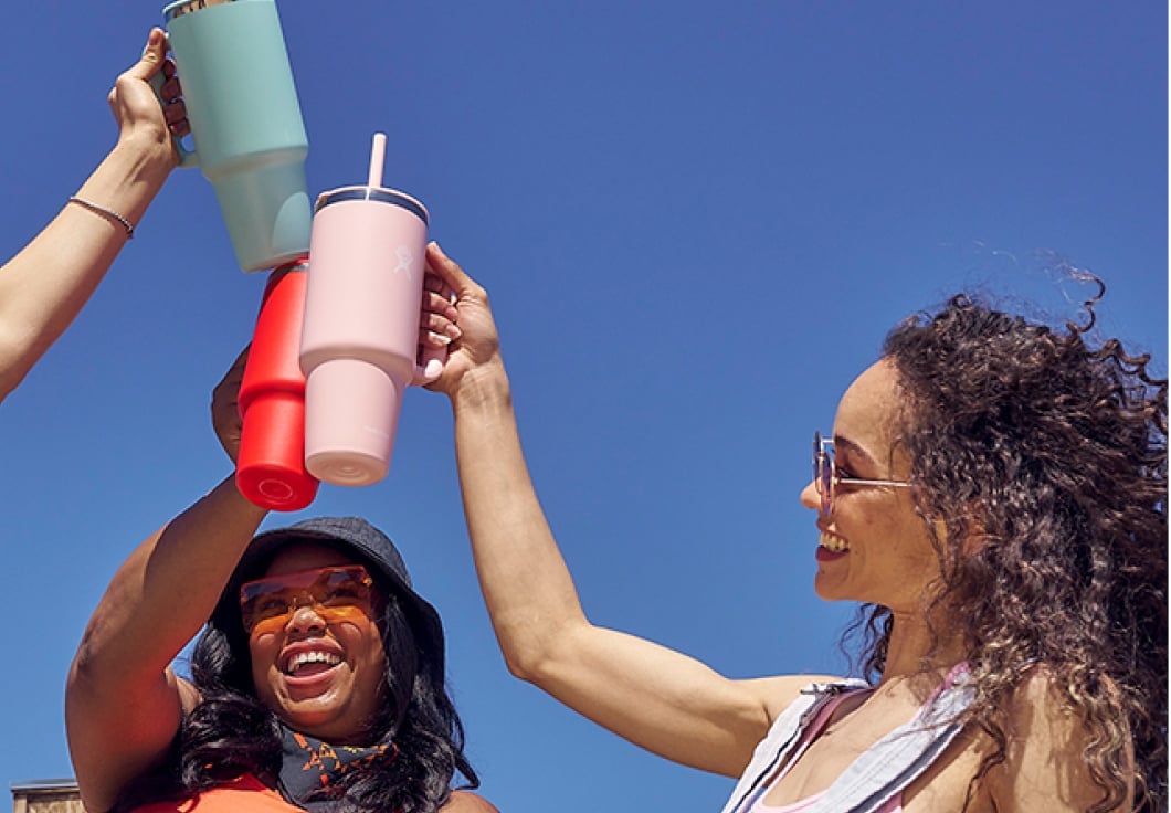 Three diverse Gen-Z females cheer their Hydro Flask Travel Tumblers in a Jeep, bodies out of the convertible top. Two girls in sunglasses, vibrant blue, red, and pink tumblers in hand, against a blue sky backdrop.