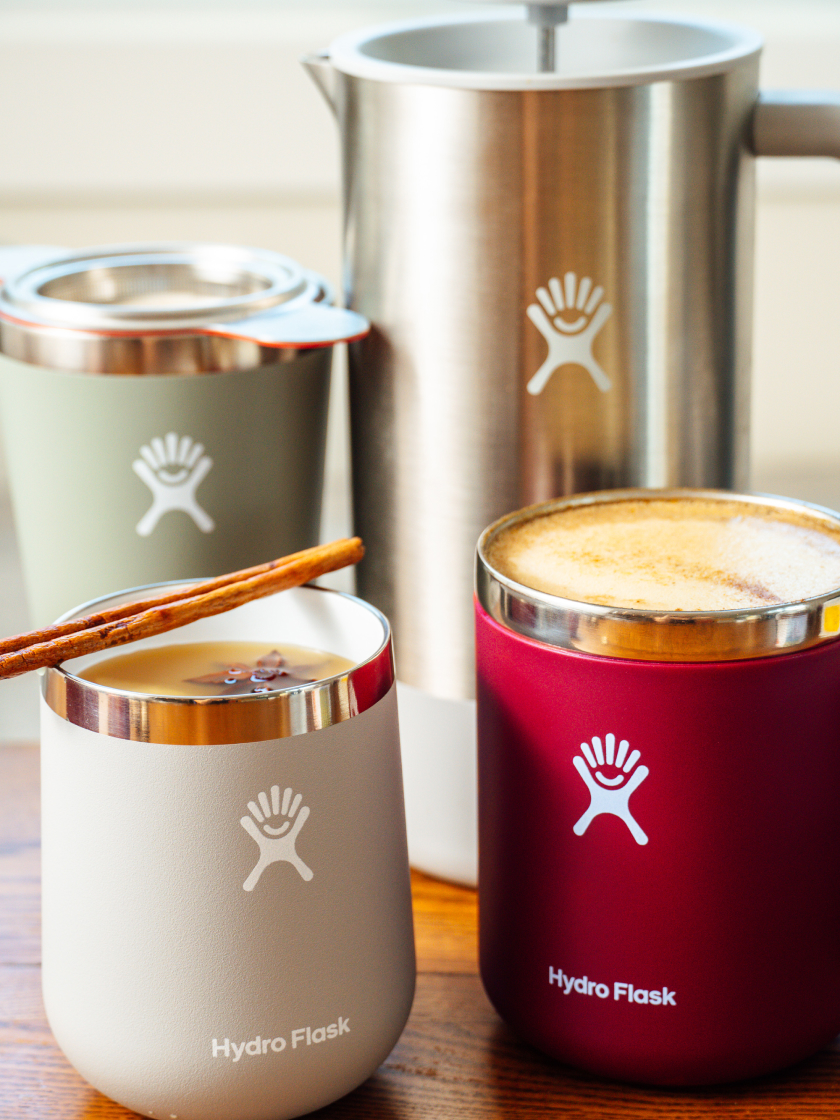 Drei köstliche Herbstgetränke auf einer hölzernen Küchenarbeitsplatte: Pumpkin Spice Latte in einem beerenfarbenen Becher, Maple Ginger Mulled Apple Cider in einem grauen Wein-Tumbler mit Zimtstange und ein London Fog Tea in einem Agave-farbenen Tumbler mit einem ... aus Edelstahl