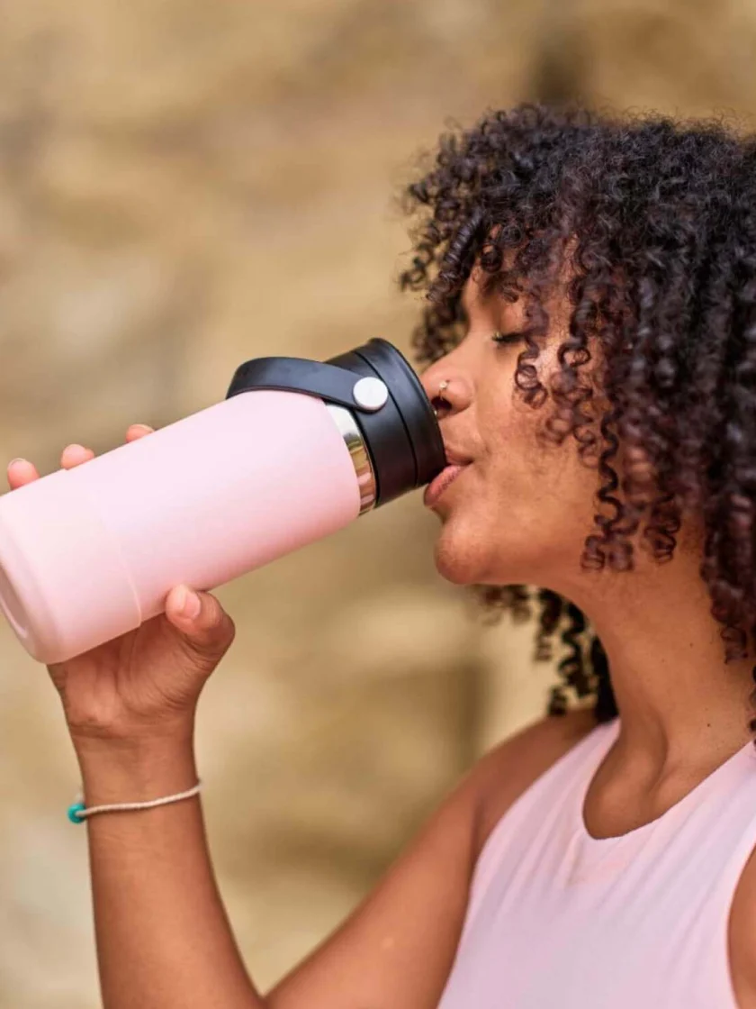 A woman with curly hair and a pink tank top is drinking from a Hydro Flask 20 oz Water Bottle with a Flex Sip Lid.