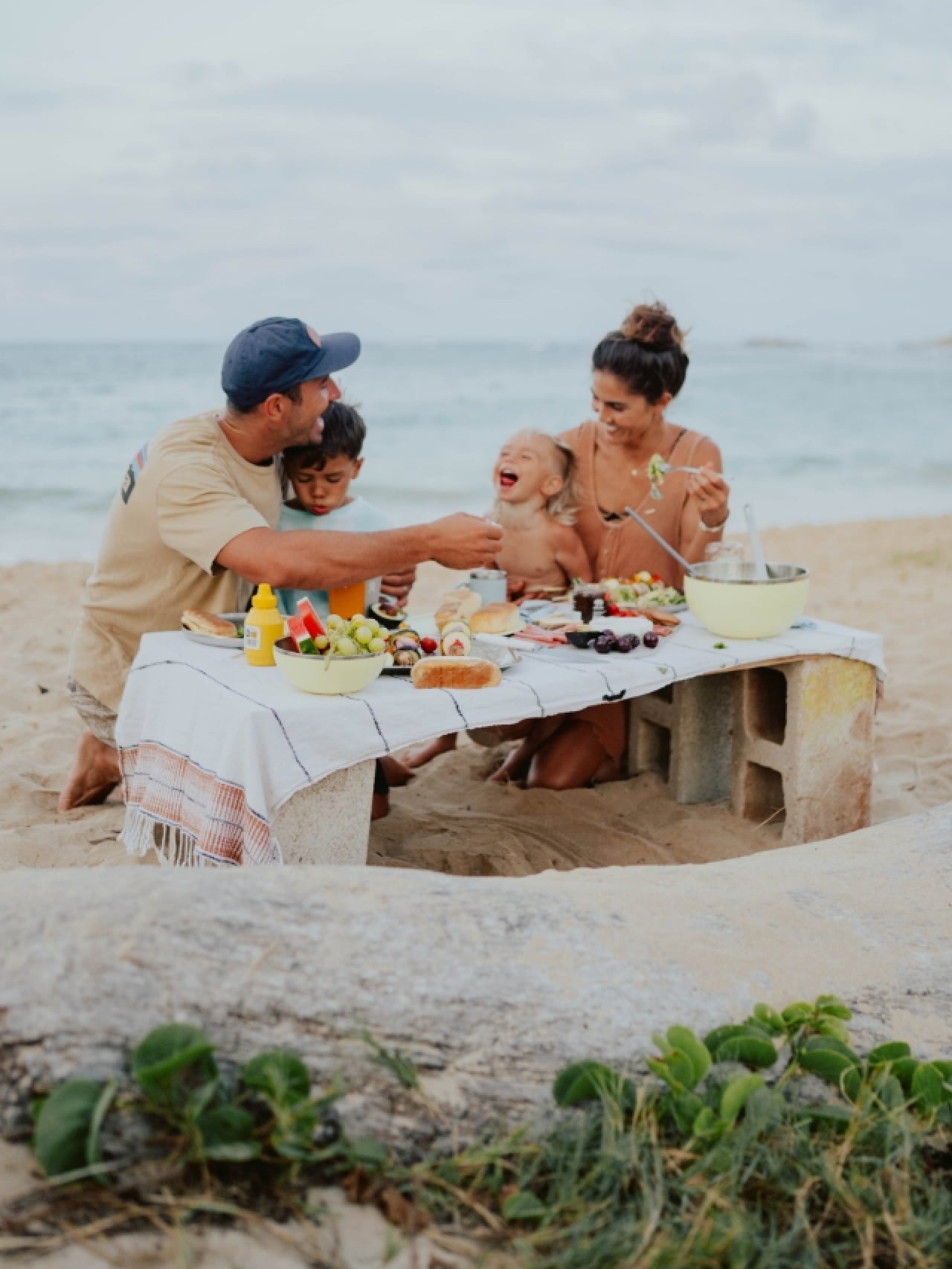 A happy family of four enjoying a beach picnic with veggie skewers and food served in Hydro Flask Outdoor Kitchen bowls and plates on a wooden table covered with a white blanket, with the waves and sand in the background. The parents, in their early 30s w